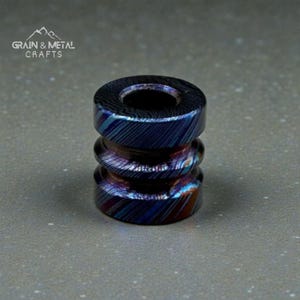 May include: A cylindrical metal bead with a textured, layered design in shades of blue, purple, and bronze. The bead has a central hole and a ridged, stacked appearance. The text "GRAIN & METAL CRAFTS" is in the upper left corner.