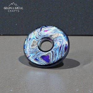 Op de afbeelding: Een ronde kraal met een centraal gat, versierd met een wervelend patroon in blauw, paars en zilver. De rand is donker. De tekst "GRAIN & METAL CRAFTS" is zichtbaar. Het ontwerp doet denken aan stromend water.