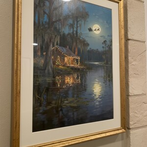 K&ouml;nnte beinhalten: Gerahmter Kunstdruck, der eine gem&uuml;tliche H&uuml;tte an einem See bei Nacht zeigt, beleuchtet von Weihnachtsbeleuchtung. Ein Vollmond scheint dar&uuml;ber, mit einer Silhouette des Weihnachtsmanns. Das Kunstwerk ist in einem goldfarbenen Rahmen.