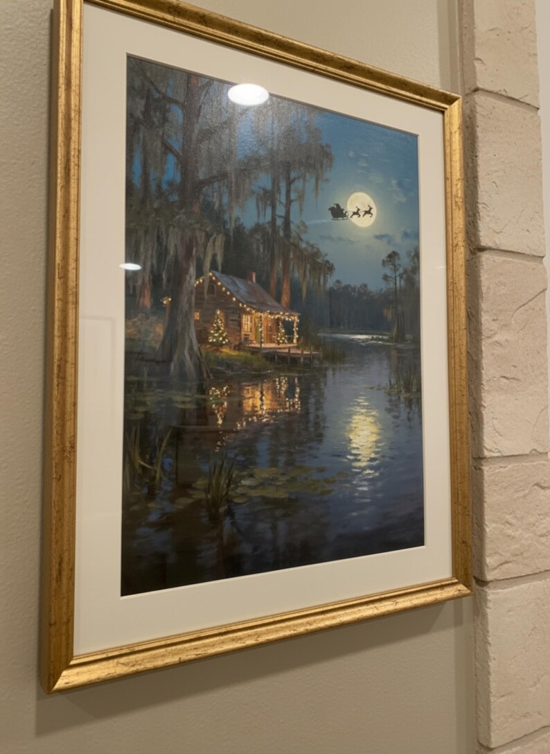K&ouml;nnte beinhalten: Gerahmter Kunstdruck, der eine gem&uuml;tliche H&uuml;tte an einem See bei Nacht zeigt, beleuchtet von Weihnachtsbeleuchtung. Ein Vollmond scheint dar&uuml;ber, mit einer Silhouette des Weihnachtsmanns. Das Kunstwerk ist in einem goldfarbenen Rahmen.