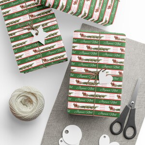 May include: Christmas-themed gift wrap featuring a repeating pattern of green crocodiles and Santa's sleigh on a white background. The words "Joyeux Noël" are printed in a festive font. The gifts are tied with twine and adorned with star-shaped tags.
