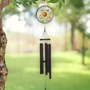 May include: A decorative wind chime with a stained glass sunflower design. The round glass panel features a yellow sunflower with green leaves and a blue butterfly. The chime has black metal tubes and a triangular clapper, hanging from a tree branch.