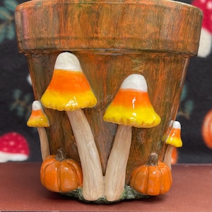 May include: A brown, textured plant pot decorated with three-dimensional sculpted mushrooms and pumpkins. The mushroom caps are painted in a candy corn pattern of white, orange, and yellow. Two small orange pumpkins sit at the base of the pot.