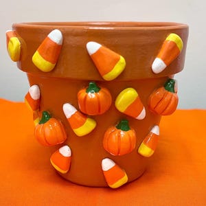 May include: A terracotta-colored plant pot decorated with three-dimensional embellishments of orange pumpkins with green stems and triangular candy corn pieces in yellow, orange, and white. The pot has a wide rim and a tapered cylindrical shape.