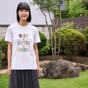 May include: A white t-shirt with the words "BEST", "BETTER", and "GOOD" in colourful, textured lettering. The words are arranged vertically with arrows pointing upwards. The person is wearing a black skirt and standing in a garden.