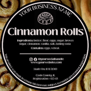 May include: Close-up of freshly baked cinnamon rolls. A black circular label displays "Cinnamon Rolls" with ingredients listed. The label also includes social media handles and website information. The rolls are golden brown with a swirl pattern.