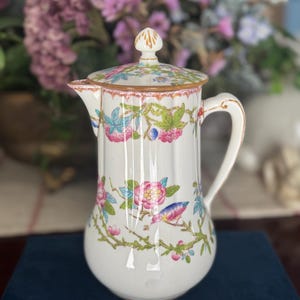 May include: White ceramic pitcher with a lid, adorned with floral and bird motifs in pink, blue, and green. The pitcher has a handle and a spout, and the lid is topped with a gold finial. The design is set against a white background.