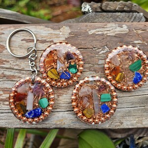 May include: Four round, copper-colored pendants with a variety of colorful gemstones and crystals. One pendant is attached to a silver keychain. The gemstones include blue, green, yellow, and orange stones. The pendants are on a wooden surface.