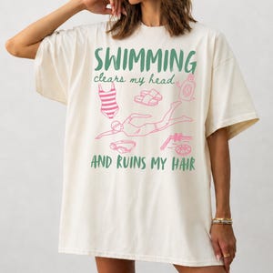 Könnte beinhalten: Ein übergroßes naturfarbenes T-Shirt mit dem grünen Text SWIMMING clears my head AND RUINS MY HAIR. Das Design enthält rosa Strichzeichnungen eines gestreiften Badeanzugs, Sandalen, einer Flasche, einer schwimmenden Person und einer Schwimmbrille.