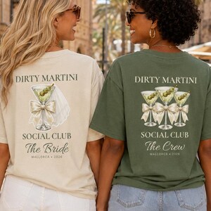 Könnte beinhalten: Ein beiges T-Shirt mit einer Martiniglas-Grafik, einer weißen Schleife und dem Text 'DIRTY MARTINI SOCIAL CLUB The Bride MALLORCA 2026' sowie ein olivgrünes T-Shirt mit drei Martinigläsern, Schleifen und 'DIRTY MARTINI SOCIAL CLUB The Crew MALLORCA 2026'.