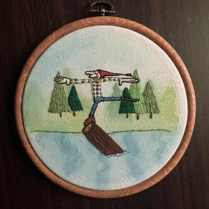 Peut inclure: Broderie dans un cercle en bois. Le motif représente un personnage de dessin animé en équilibre sur une bûche dans l'eau, avec des arbres en arrière-plan. Le personnage porte un chapeau rouge, une chemise à carreaux et un jean bleu. L'œuvre est encadrée dans un cercle en bois.