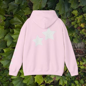 Pode incluir: Um moletom rosa claro com capuz e mangas compridas. A parte de trás do moletom apresenta duas grandes estrelas brancas com um padrão cinza claro. A manga esquerda tem três estrelas brancas. O moletom é feito de um material macio.