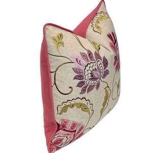 May include: Decorative throw pillow with a floral design. The pillow features a beige background with embroidered flowers in shades of purple, pink, and green. The edges and back of the pillow are a solid pink color.