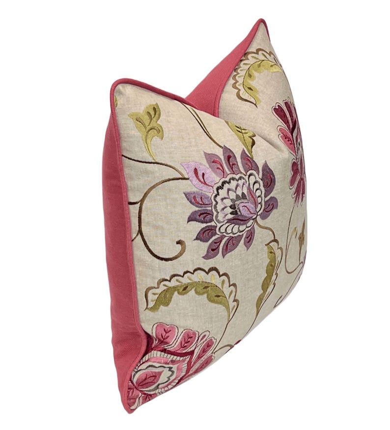 May include: Decorative throw pillow with a floral design. The pillow features a beige background with embroidered flowers in shades of purple, pink, and green. The edges and back of the pillow are a solid pink color.