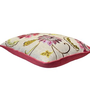 May include: Square decorative pillow with a floral pattern. The pillow features a light beige background with embroidered flowers in shades of pink, purple, and green. The edges are trimmed with a solid pink border.