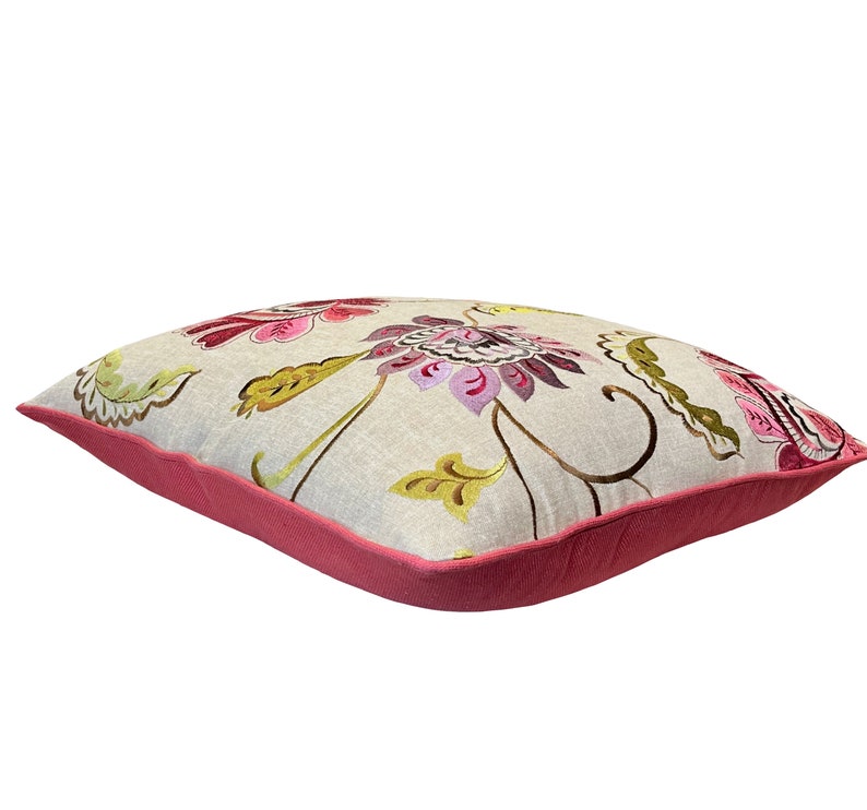 May include: Square decorative pillow with a floral pattern. The pillow features a light beige background with embroidered flowers in shades of pink, purple, and green. The edges are trimmed with a solid pink border.