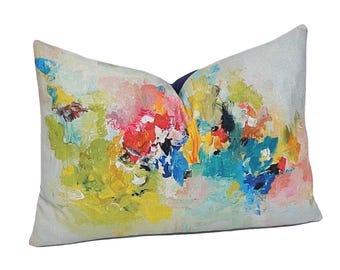 Könnte beinhalten: Ein dekoratives rechteckiges Kissen mit einem abstrakten, gemalten Design. Das Kunstwerk zeigt eine Mischung aus leuchtenden Farben, darunter Gelb, Rot, Blau und Grün, die einen strukturierten, künstlerischen Eindruck erzeugen. Das Kissen hat einen hellen Hintergrund.
