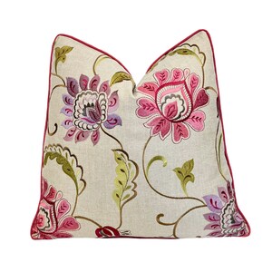 May include: Decorative throw pillow with a floral pattern. The pillow features embroidered flowers in shades of pink, purple, and green on a cream-colored background. A dark red trim outlines the pillow's edges, adding a contrasting detail.