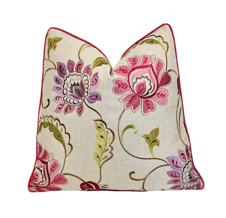 May include: Decorative throw pillow with a floral pattern. The pillow features embroidered flowers in shades of pink, purple, and green on a cream-colored background. A dark red trim outlines the pillow's edges, adding a contrasting detail.