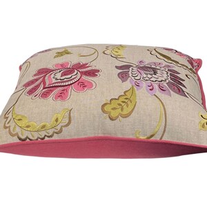 May include: Square decorative pillow with a floral pattern. The pillow features a beige background with large pink and purple flowers, green leaves, and a pink border. The pillow is a square shape.