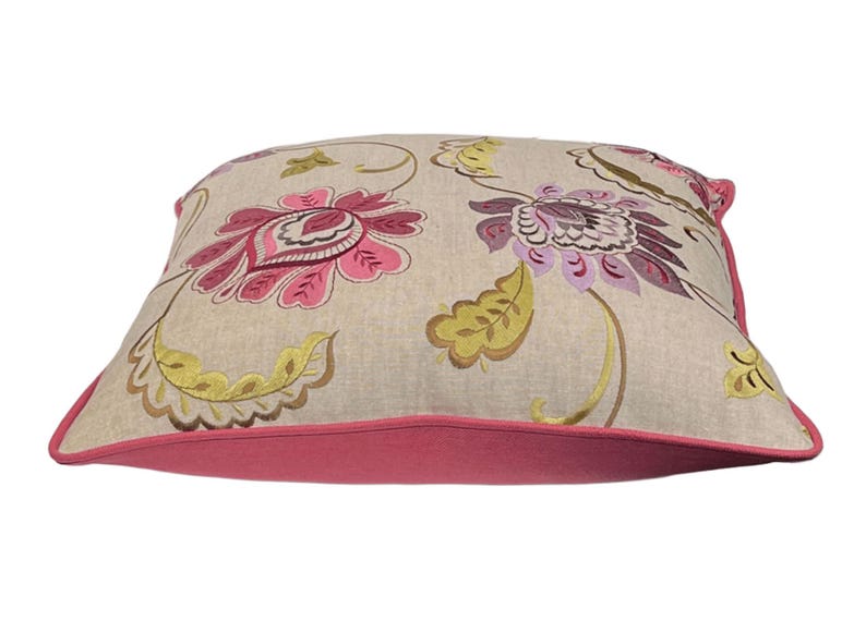 May include: Square decorative pillow with a floral pattern. The pillow features a beige background with large pink and purple flowers, green leaves, and a pink border. The pillow is a square shape.