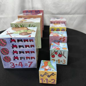 May include: A collection of colorful, hand-painted wooden blocks. Each block features unique illustrations, including trains, apples, and musical notes, along with simple math problems. The blocks are arranged in a staggered display, showcasing the variety of designs.