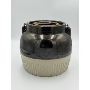 May include: A two-toned ceramic crock featuring a dark brown glazed upper section and a light beige, vertically ribbed lower base. The vessel includes two small side handles and a matching circular lid nested within the top opening.
