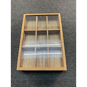 May include: A rectangular wooden display box featuring a clear glass lid and nine individual compartments arranged in a three-by-three grid. The frame is constructed from light brown wood with a visible grain, providing a structured storage solution.
