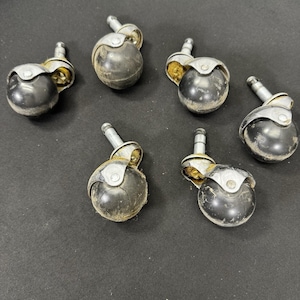 May include: Six antique black and silver furniture casters. Each caster has a black, spherical wheel, a silver metal housing, and a silver stem for insertion. The casters show signs of age and wear, with some dust and discoloration.
