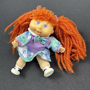 May include: A vintage doll with orange yarn hair styled in pigtails. The doll has blue eyes, a peach-colored face, and is wearing a purple and floral dress with a green bow. The doll's shoes are white with black accents.