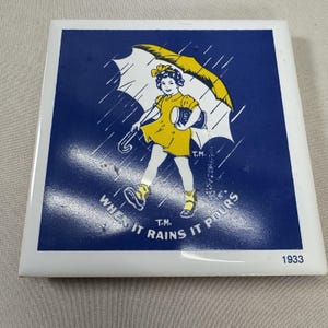 May include: A square ceramic tile with a blue background and a white border. The tile features a vintage illustration of a girl in a yellow dress and boots holding an umbrella. The text "WHEN IT RAINS IT POURS" is also on the tile, along with the year 1933.