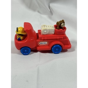 May include: A red Little Tikes fire truck toy with blue wheels. The toy truck features a firefighter figure in a yellow helmet and a brown dog figure. The toy has the words "Little Tikes" printed on the side.