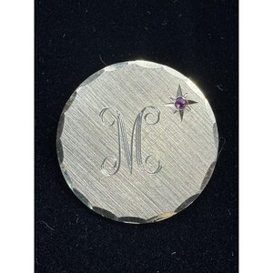 May include: A circular silver-toned metal disc featuring a brushed finish and a scalloped edge. The center is engraved with a large, cursive letter M. A small, four-pointed star design is etched to the right of the letter, set with a tiny pink stone.