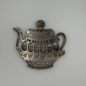 May include: A silver-colored metal teapot-shaped trivet. The trivet features an openwork design with seashell motifs and decorative detailing. The handle and spout are curved. The lid is topped with a seashell. The trivet is designed to protect surfaces from hot teapots.