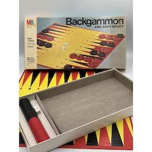 May include: A Milton Bradley Backgammon and Acey-Deucey board game set featuring a yellow and red board with black triangular points. The box lid displays the game title, while the open box contains a red and black dice cup and two white dice.