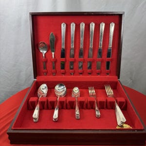 May include: A silver-toned flatware set arranged in a dark wooden storage case with a vibrant red velvet interior. The set includes various spoons, forks, and knives held in individual slots, featuring a polished metallic finish and simple handles.