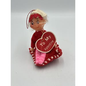 May include: A small doll with white hair, wearing a red outfit and a white bonnet, holding a red heart-shaped tag that reads To My Valentine. The doll has pink and white striped legs and is seated on a red fabric base with white trim.