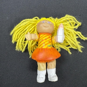 May include: A vintage Cabbage Patch Kids doll with yellow yarn hair, wearing an orange and yellow dress, white socks, and shoes. The doll is holding a white and pink lipstick. The doll is on a black surface.