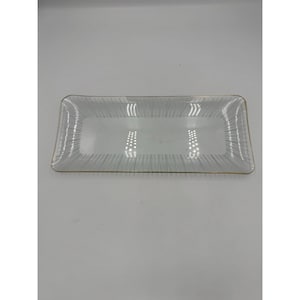 May include: A rectangular clear glass tray featuring a delicate pattern of thin, vertical white lines radiating from the center toward the edges. The tray is finished with a thin, metallic gold-colored rim along the entire outer perimeter.