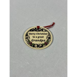 May include: Oval Christmas ornament with the text "Merry Christmas to a great Grandpa." The ornament has a cream-colored background with a black border and holly berry accents. A red ribbon is attached for hanging.