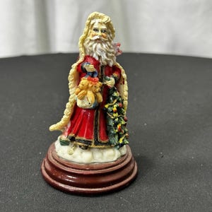 May include: A small Santa Claus figurine, dressed in a red coat with white trim and a long white beard. He holds a small Christmas tree and a bag with teddy bears. The figurine is mounted on a round, brown base.