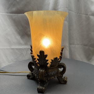 May include: A decorative table lamp with a warm, amber-colored glass shade. The lamp has a bronze-colored base with ornate, leaf-like details. The lamp is lit, emitting a soft glow. The lamp's cord is visible.