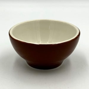 May include: A small, round ceramic bowl featuring a glossy, deep brown exterior and a smooth, off-white interior. The bowl has a simple, curved silhouette with a slightly tapered base, providing a clean and functional design for serving food.