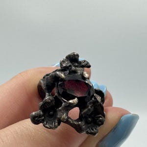 May include: A dark silver-toned ring featuring an oval-shaped, deep red faceted gemstone set within a textured, organic metal frame that resembles intertwined floral or botanical elements with a weathered, oxidized finish and intricate detailing.