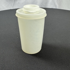 May include: A translucent, off-white plastic container with a lid. The container is cylindrical with a slightly flared base. The lid has a raised, circular center and a flat rim. The container appears to be empty.