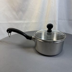 May include: A stainless steel saucepan with a black handle and a clear glass lid. The handle has a loop for hanging. The lid has a black knob. The pan is sitting on a gray surface.