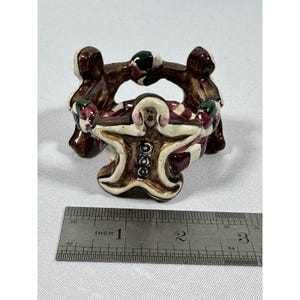May include: A ceramic gingerbread man candle holder. The holder is brown, white, and red with green accents. The gingerbread man is in the center, surrounded by three figures. The candle holder measures approximately 3 inches in length.