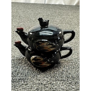 May include: A stackable black ceramic teapot set featuring two individual pieces shaped like roosters. Each piece has a glossy black finish, a red comb on the head, and a handle on the side. The top piece includes a lid with a small black bird finial.