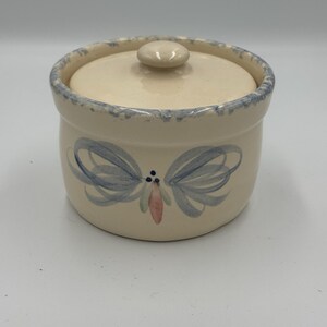 May include: A cream-colored ceramic lidded jar with a cylindrical body and a matching lid. The rim features a blue speckled pattern, and the front is decorated with a hand-painted blue and pink floral motif consisting of looped petals and dots.