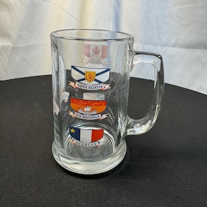 May include: Clear glass beer mug with a handle. The mug features colorful emblems of Canadian provinces, including Nova Scotia, New Brunswick, and Acadian. The mug has a wide base and a cylindrical body.